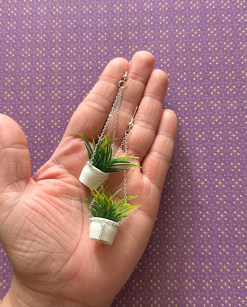 May include: A pair of silver dangle earrings featuring miniature white hanging planters with green plants.