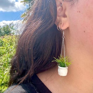 May include: A pair of dangle earrings featuring a small white pot with a green plant hanging from a silver chain.