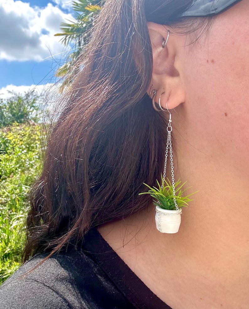May include: A pair of dangle earrings featuring a small white pot with a green plant hanging from a silver chain.