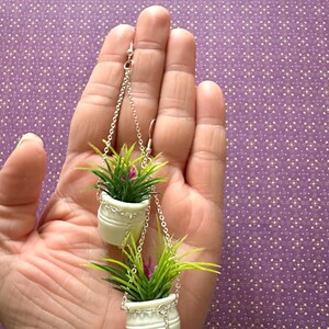 May include: A pair of dangle earrings featuring two white ceramic pots with green plants hanging from silver chains.