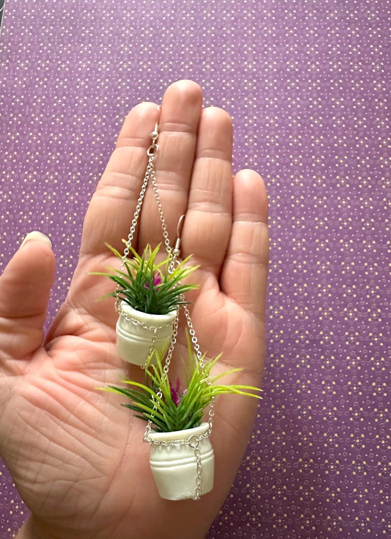 May include: A pair of dangle earrings featuring two white ceramic pots with green plants hanging from silver chains.