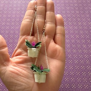 May include: A pair of dangle earrings featuring miniature hanging planters with green leaves and pink flowers. The planters are white and have a silver chain.