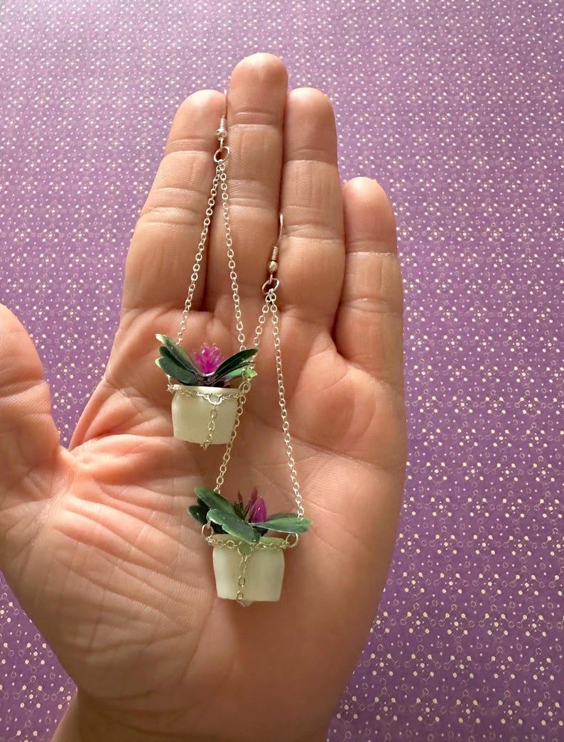 May include: A pair of dangle earrings featuring miniature hanging planters with green leaves and pink flowers. The planters are white and have a silver chain.