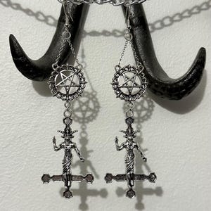 May include: A pair of silver earrings featuring a pentagram charm and a figure of a demon on a cross pendant.