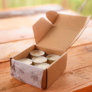 Puede incluir: Una caja de cartón con la tapa abierta, que revela seis pequeñas velas de té redondas y blancas. La caja está forrada con papel de seda blanco con un diseño floral.