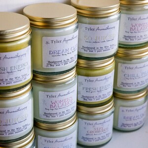 May include: A collection of glass candle jars with gold lids. The jars are filled with white wax and have labels with different names and descriptions. The labels include "So Juiced", "Dream Cloud", "Women's Balance", "Chill Pill", "Fresh Energy", "Stay Grounded".