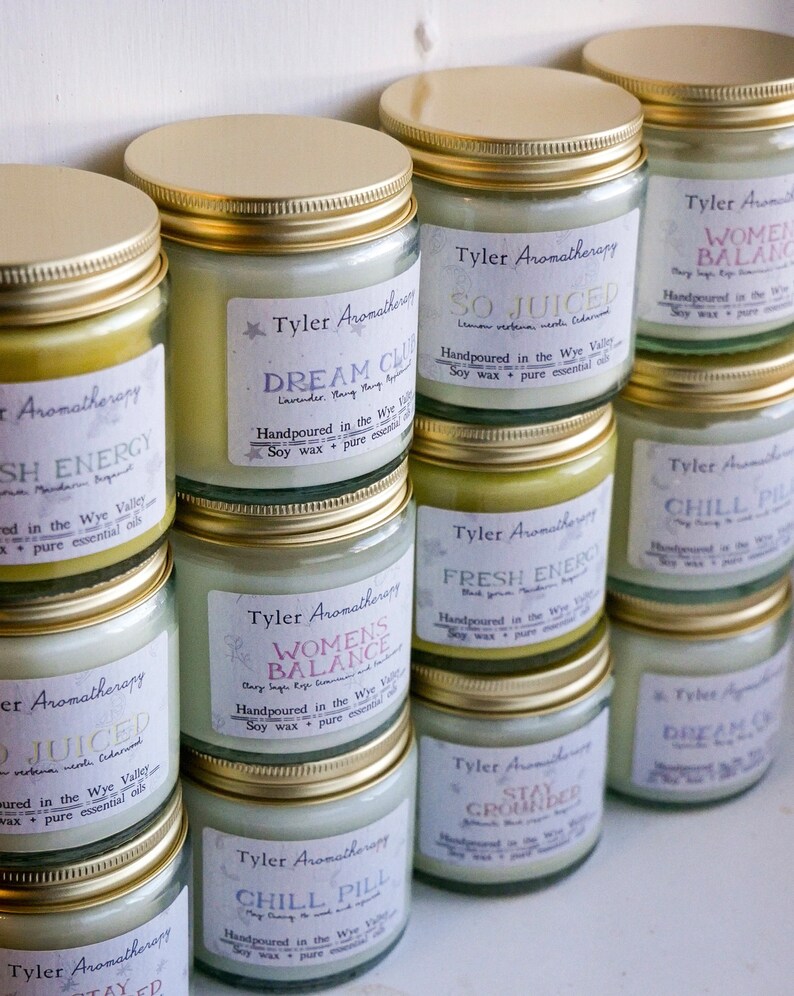 May include: A collection of glass candle jars with gold lids. The jars are filled with white wax and have labels with different names and descriptions. The labels include "So Juiced", "Dream Cloud", "Women's Balance", "Chill Pill", "Fresh Energy", "Stay Grounded".
