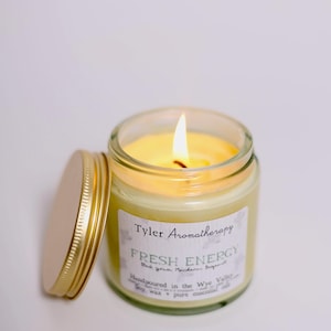 May include: A lit candle in a glass jar with a gold lid. The label reads "Tyler Aromatherapy Fresh Energy Blackcurrant, Mandarin, Bergamot Handpoured in the Wye Valley Soy wax + pure essential oils".