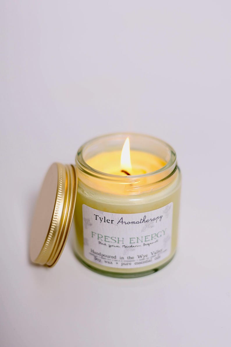 May include: A lit candle in a glass jar with a gold lid. The label reads "Tyler Aromatherapy Fresh Energy Blackcurrant, Mandarin, Bergamot Handpoured in the Wye Valley Soy wax + pure essential oils".