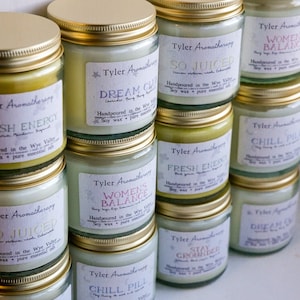 Può includere: Una collezione di barattoli di vetro per candele con coperchi dorati. I barattoli sono riempiti con candele di cera di soia e hanno etichette con diversi profumi e nomi, come "Fresh Energy", "Dream Cloud", "So Juiced", "Women's Balance", "Chill Pill" e "Stay Grounded".
