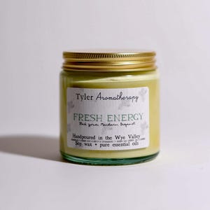 May include: A glass jar candle with a gold lid and a white label that reads "Tyler Aromatherapy Fresh Energy Black Spruce, Mandarin, Bergamot Handpoured in the Wye Valley Soy wax + pure essential oils"