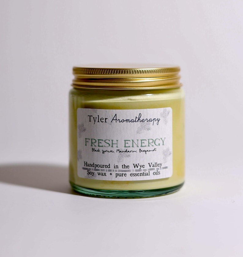 May include: A glass jar candle with a gold lid and a white label that reads "Tyler Aromatherapy Fresh Energy Black Spruce, Mandarin, Bergamot Handpoured in the Wye Valley Soy wax + pure essential oils"
