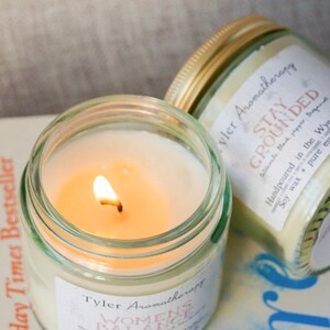 May include: Two glass candle jars with gold lids. The candle on the left is lit and has a label that reads "Tyler Aromatherapy Women's Balance Handpoured in the Wye Soy wax + pure essential oils". The candle on the right has a label that reads "Tyler Aromatherapy Stay Grounded Handpoured in the Wye Soy wax + pure essential oils"