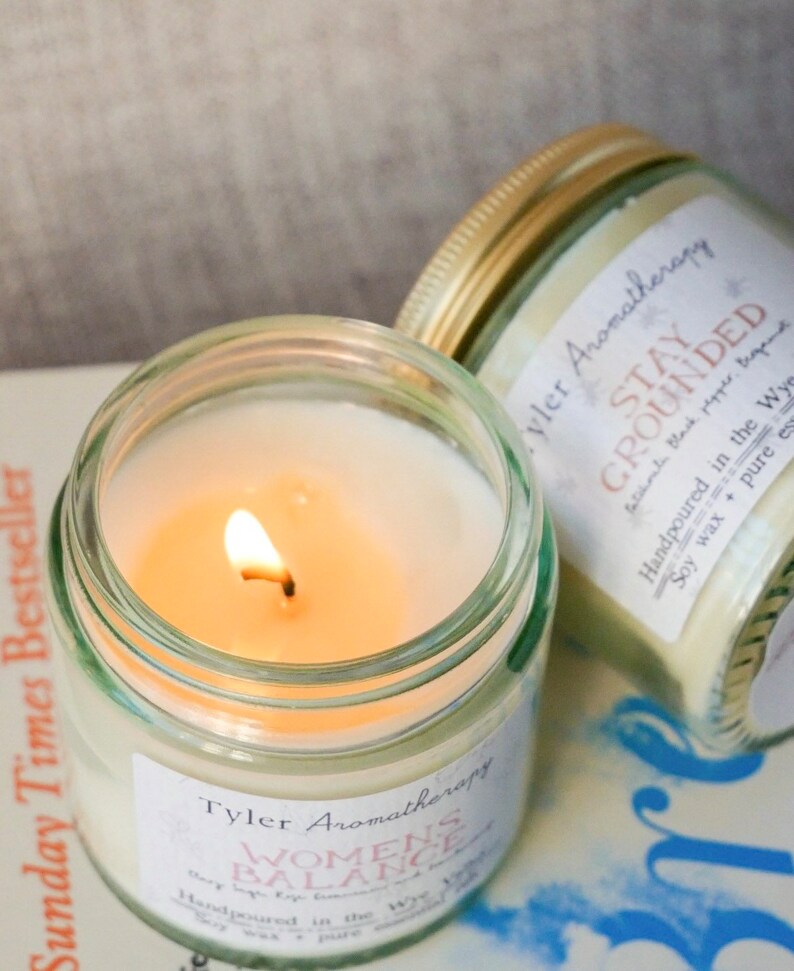 May include: Two glass candle jars with gold lids. The candle on the left is lit and has a label that reads "Tyler Aromatherapy Women's Balance Handpoured in the Wye Soy wax + pure essential oils". The candle on the right has a label that reads "Tyler Aromatherapy Stay Grounded Handpoured in the Wye Soy wax + pure essential oils"