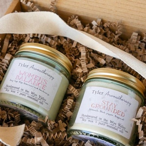 May include: Two glass candle jars with gold lids and labels. The labels are white with black text. The left jar has a label that reads "Women's Balance" with the text "Clary Sage, Rose, Geranium and Frankincense" below. The right jar has a label that reads "Stay Grounded" with the text "Patchouli, Black Pepper, Bergamot" below. Both labels have the text "Handpoured in the Wye Valley" and "Soy wax + pure essential oils" below the scent descriptions.