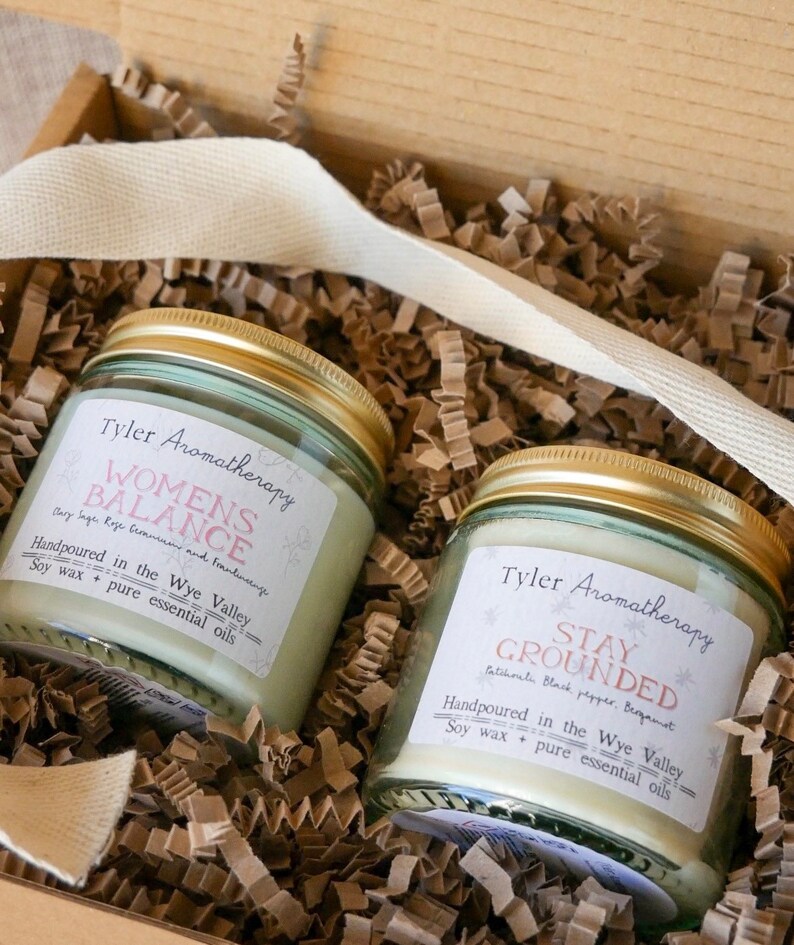 May include: Two glass candle jars with gold lids and labels. The labels are white with black text. The left jar has a label that reads "Women's Balance" with the text "Clary Sage, Rose, Geranium and Frankincense" below. The right jar has a label that reads "Stay Grounded" with the text "Patchouli, Black Pepper, Bergamot" below. Both labels have the text "Handpoured in the Wye Valley" and "Soy wax + pure essential oils" below the scent descriptions.