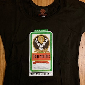 May include: Black t-shirt with a green and white Jägermeister logo featuring a stag with antlers and a cross. The text on the logo reads "Jägermeister Auszug Edelster Kräuter" and "Kräuter-Liqueur". The text "IMPORTED" is at the top of the logo. The text "SERVE COLD - KEEP ON ICE" is at the bottom of the logo.