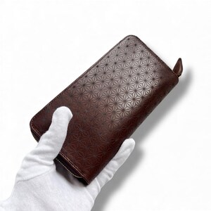 Puede incluir: Cartera de cuero marrón con un patrón geométrico de estrellas. La cartera es rectangular con cierre de cremallera y una pequeña lengüeta de cuero. La cartera es sostenida por una mano enguantada sobre un fondo blanco.