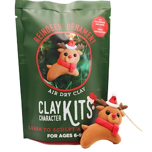 Peut inclure: Un paquet vert contenant un kit d'ornement de renne. Le paquet présente un renne de dessin animé avec une écharpe rouge et un bonnet de Père Noël. Le texte sur le paquet indique "Reindeer Ornament", "Air Dry Clay" et "Clay Kits". Le kit est pour les enfants de 6 à 9 ans.
