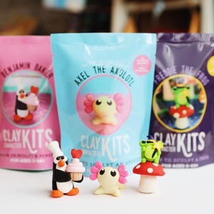 Könnte beinhalten: Drei bunte Pakete mit Ton-Sets für Kinder. Jedes Paket zeigt ein anderes Tier: einen Pinguin, einen Axolotl und einen Frosch. Die Pakete sind rosa, blau und lila und tragen den Schriftzug "Clay Kits".