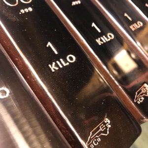 May include: Close-up of several copper bars. Each bar is stamped with "CU .999" and "1 KILO". The bars have a reflective surface and a copper color. A logo is visible on the bottom right of each bar.
