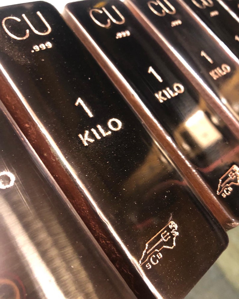 May include: Close-up of several copper bars. Each bar is stamped with "CU .999" and "1 KILO". The bars have a reflective surface and a copper color. A logo is visible on the bottom right of each bar.
