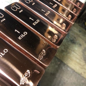 May include: Close-up of several copper bars. Each bar is stamped with "CU .999" and "1 KILO". The bars have a shiny, metallic finish and a small logo is visible on each bar. The bars are arranged in a row.