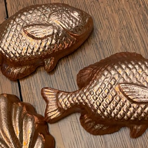 May include: Three bronze-colored fish-shaped decorative objects. The fish have detailed scale patterns and fins. The objects are arranged on a wooden surface, suggesting a nautical or coastal theme.