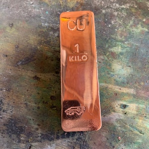 May include: A copper bar with the stamped text "CU" and "1 KILO". The bar has a shiny, reflective surface and a logo at the bottom. The bar is a reddish-brown color.