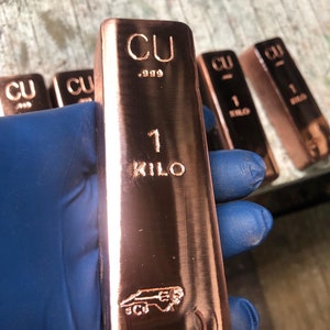 May include: A copper bar with the markings "CU .999" and "1 KILO" is held by a gloved hand. The bar has a shiny, reflective surface and a logo at the bottom. Several similar bars are visible in the background.
