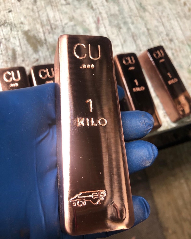 May include: A copper bar with the markings "CU .999" and "1 KILO" is held by a gloved hand. The bar has a shiny, reflective surface and a logo at the bottom. Several similar bars are visible in the background.