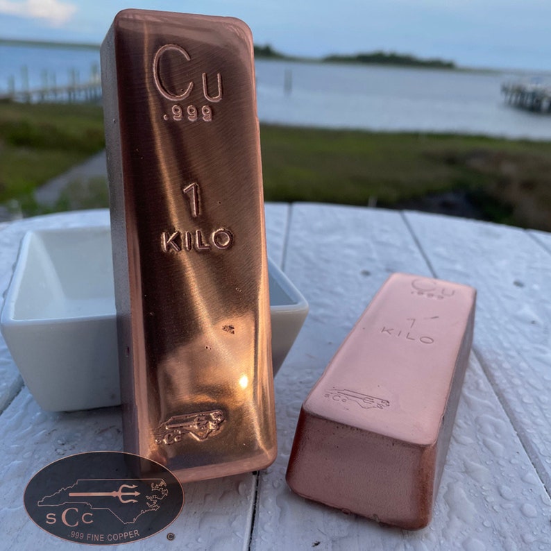 May include: Two copper bars, each weighing 1 kilo, are displayed. The bars are stamped with "Cu .999" and "1 KILO". A small oval emblem with text and a trident is visible. The bars have a shiny, metallic finish.