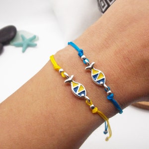Könnte beinhalten: Zwei Freundschaftsarmbänder mit verstellbaren Knoten. Ein Armband ist blau mit einem silbernen Fisch-Anhänger mit gelber und blauer Emaille. Das andere Armband ist gelb mit einem silbernen Fisch-Anhänger mit gelber und blauer Emaille.