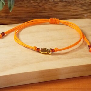 May include: An adjustable orange cord bracelet featuring a central gold-toned fish charm. The bracelet includes small dark red beads on each side of the fish and at the ends of the cord, with a sliding knot mechanism for size adjustment.
