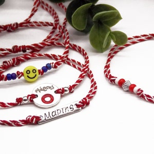Op de afbeelding: Drie rode en witte touw armbanden met zilveren bedels. Een bedel heeft een smiley, een andere zegt "μάγεια" en heeft een rode cirkel, en de derde zegt "Μαρία"
