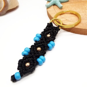 May include: Black and turquoise blue macrame keychain with a gold key ring. The keychain has small gold beads interspersed throughout the macrame.