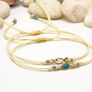 Puede incluir: Dos pulseras delicadas con dijes de peces blancos y azules. Las pulseras están hechas de cordón crema y tienen pequeñas cuentas azules.