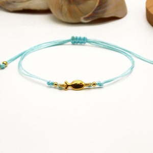 Könnte beinhalten: Ein hellblaues, verstellbares Armband mit einem goldfarbenen Fischanhänger. Das Armband besteht aus dünnem, geflochtenem Kordel mit goldfarbenen Perlen an jedem Ende. Der Fischanhänger befindet sich in der Mitte des Armbands.