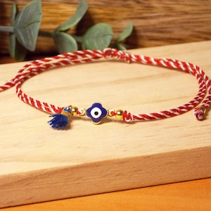 Peut inclure: Un bracelet tressé rouge et blanc avec un charme en forme d'œil bleu et doré et un petit pompon bleu. Le bracelet est présenté sur une surface en bois, avec de la verdure en arrière-plan.