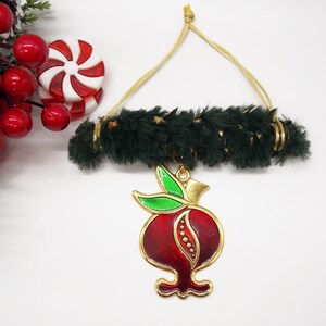 May include: A gold-coloured ornament shaped like a pomegranate with red enamel and green leaves. The ornament is hanging from a green and gold fuzzy garland.