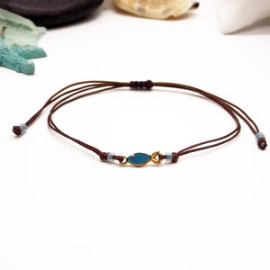 May include: A brown cord bracelet with a small gold fish charm. The fish is blue with a gold outline.