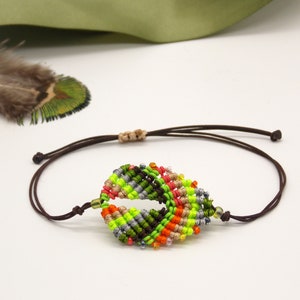 Puede incluir: Un brazalete de hoja de color hecho con cordones anudados en tonos de verde, naranja, rojo y marrón. El brazalete está adornado con cuentas plateadas y transparentes y tiene un cierre de cordón marrón.