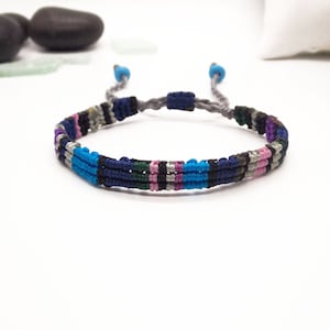 Puede incluir: Un brazalete trenzado gris con un patrón multicolor de hilos azules, rosas, negros y verdes.