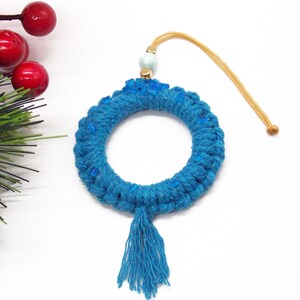 May include: A teal blue macrame wreath ornament with a tassel and blue glass beads. The wreath has a gold cord for hanging.