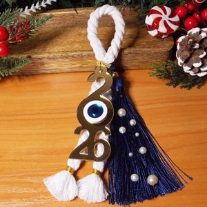 May include: A gold-colored charm with the number 2020, featuring an evil eye design. It has a white rope loop, white tassels, and a navy blue tassel with faux pearls. The charm is set against a wooden background with holiday decorations.