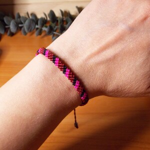 Puede incluir: Pulsera de amistad hecha a mano en tonos rosa, rojo y negro, tejida con un patrón de chevrón. La pulsera está en una muñeca, mostrando el intrincado anudado y la combinación de colores. El fondo es una superficie de madera.
