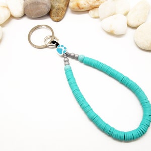 May include: A turquoise blue beaded keychain with a silver paw print charm and a silver key ring. The keychain is made of round beads and is strung on a black cord.