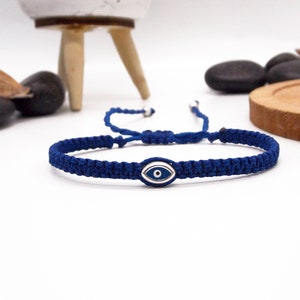 Peut inclure: Bracelet en macramé bleu marine orné d'un charme central argenté en forme d'œil protecteur. Le bracelet est réglable grâce à un fermoir coulissant. Fond blanc avec éléments décoratifs.