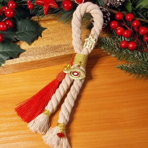 May include: A decorative hanging ornament made of thick beige twisted rope, featuring a bright red tassel, a gold-toned eye charm with a red center, and a small gold house-shaped charm labeled 2024, accented with gold thread and beads.