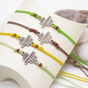 Pode incluir: Quatro pulseiras com um amuleto de cacto prateado e contas marrons. As pulseiras são de cor verde, marrom, amarelo e verde claro.